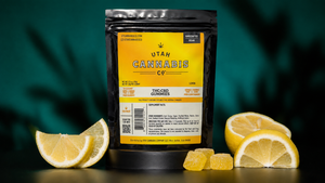 A black stand-up pouch of Utah Cannabis Co. THC-CBD Gummies with a yellow label, styled with lemon slices and three yellow gummy candies on a dark teal background.