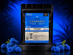 A black stand-up pouch of Utah Cannabis Co. CBD Gummies (Blue Raspberry) displayed on a black pedestal, surrounded by fresh blue raspberries on a reflective dark surface against a dark blue background with leaf shadows.