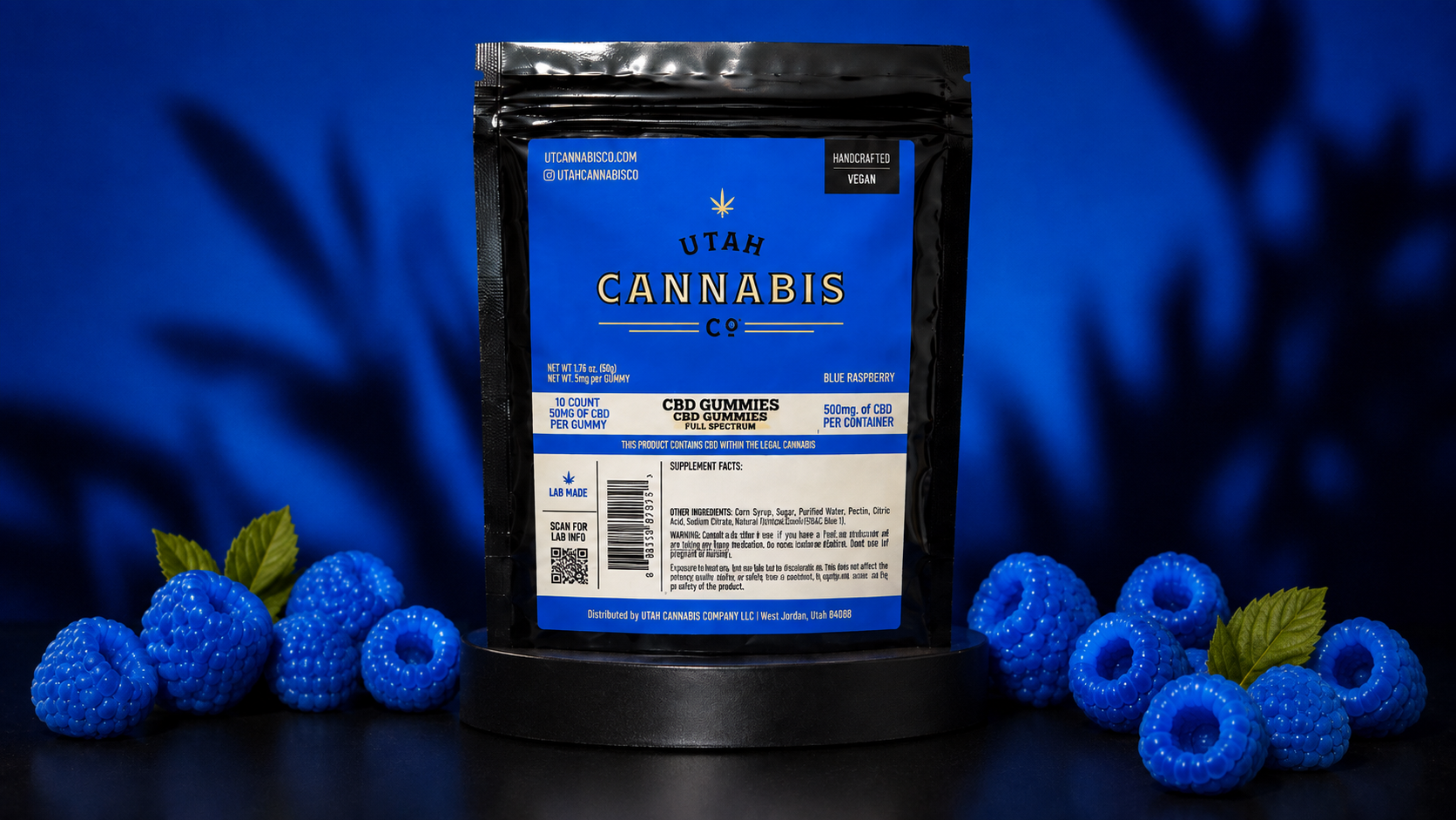 A black stand-up pouch of Utah Cannabis Co. CBD Gummies with a blue label, displayed on a dark surface with blue raspberries and green leaves around it against a blue background.