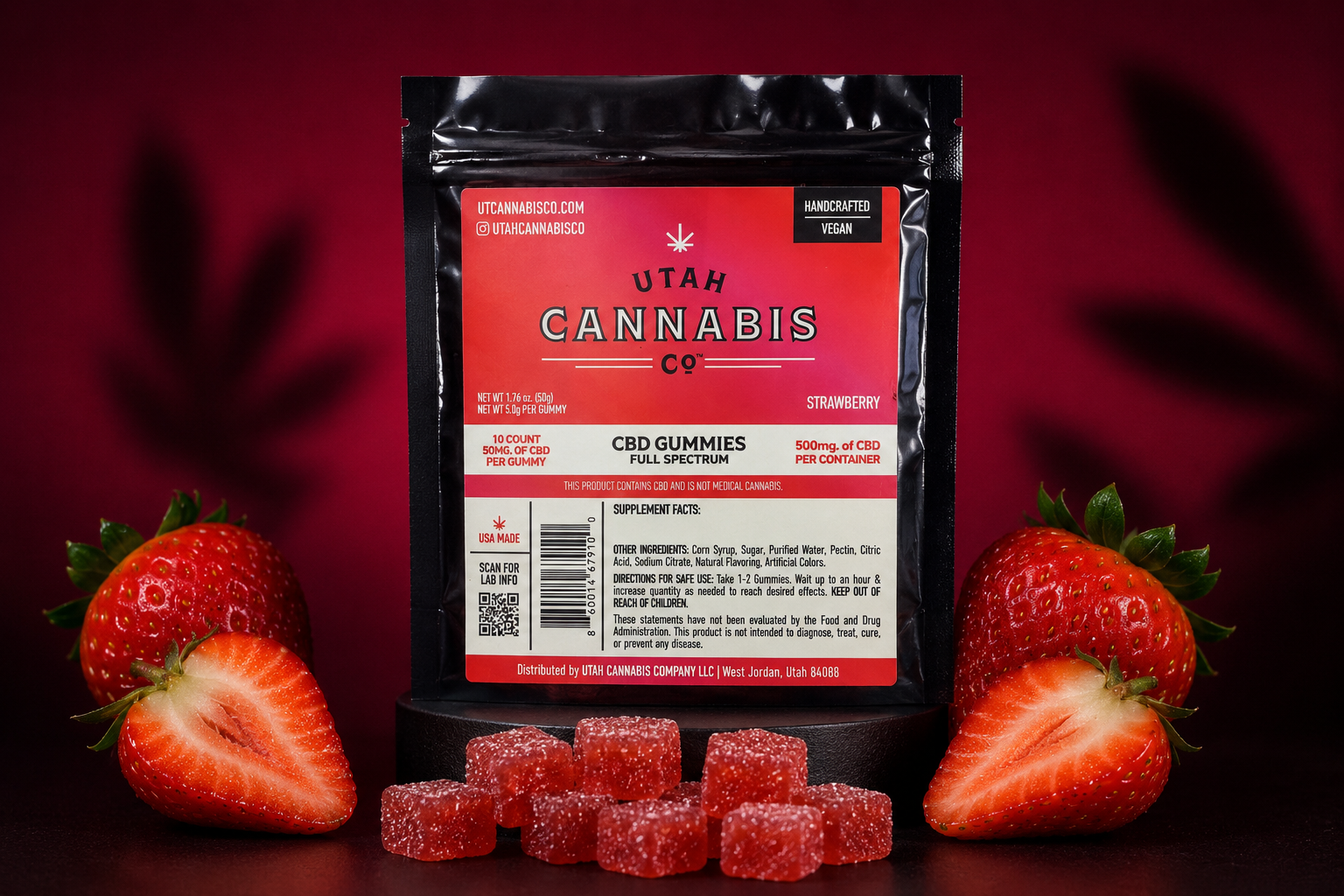 A black pouch of Utah Cannabis CV CBD Gummies, strawberry flavor, displayed on a black stand with several red gummy candies in front and fresh whole and sliced strawberries on both sides against a dark red background.