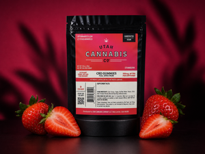 A black stand-up pouch of Utah Cannabis Co. CBD Gummies (Strawberry flavor) is centered against a dark red background with blurred strawberry shadows.